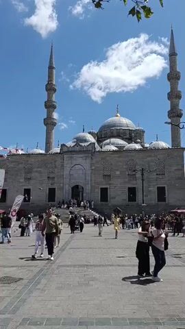 The video shows a wide shot of a large stone mosque with two tall minarets, a dome, and a central entrance with many steps leading up to it. The sky is blue with scattered white clouds. In front of the mosque, there is a large paved courtyard where many people are walking around. Some people are walking towards the mosque, while others are walking away. The people are dressed in casual summer clothing. There are also some trees and a lamppost visible in the foreground. The overall impression is a busy public space on a sunny day.
