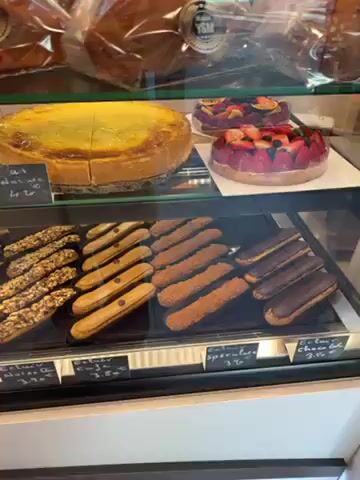 The video shows the inside of a pastry shop display case. A variety of pastries are visible, including cakes, tarts, eclairs, and individual-sized desserts. The pastries are arranged neatly on shelves within the display case. Some of the pastries have price tags with descriptions in French. A person wearing a white coat is seen briefly near the display case. The environment appears to be a brightly lit retail space.