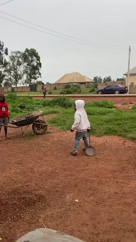 A young child wearing a white hooded sweatshirt and blue jeans is standing on a dirt path. In front of the child is a wheelbarrow full of dirt. To the left of the wheelbarrow, another child wearing a red shirt and black shorts is partially visible. In the background, there is a road with a blue car driving on it. There are also buildings and trees. The child in the white hoodie is holding a shovel and appears to be playing.