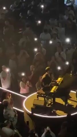 A live music performance is taking place in a crowded venue. A male musician, seated at a piano on a circular stage, is singing and playing. The stage is illuminated by yellow spotlights. The audience surrounds the stage on all sides and is using their phone lights to illuminate the space, creating a sea of light. A large screen above the stage projects a close-up of the musician as he performs. The environment is dark, except for the stage lights and the audience's phone lights. There's a sense of excitement and engagement in the crowd.