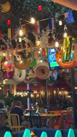 The video captures a vibrant and festive outdoor restaurant setting at night. The ceiling is covered in lush green foliage, adorned with numerous strings of globe lights that create a warm and inviting ambiance. Interspersed among the lights are a variety of national flags, suggesting a theme of international celebration or a sports-watching venue. Large inflatable rings, some resembling inner tubes, are also suspended from the ceiling, adding a playful touch. In the background, a television screen displays what appears to be a sports game. People are seated at tables, engaged in dining and conversation, their faces dimly lit by the surrounding lights. The overall atmosphere is lively and social, with a sense of outdoor dining and communal enjoyment. The camera pans slowly, offering a panoramic view of the decorated space and its patrons.