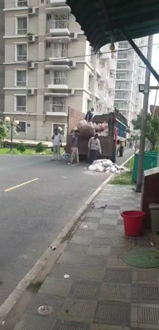 This is a video of garbage collection. A truck is parked on the side of a road, and several people are loading trash into it. The truck bed is partially filled with garbage bags. The people are wearing casual clothing. The area appears to be a residential or urban environment, with a large apartment building in the background. A sidewalk with paving stones runs along the road. There are trash cans visible near the sidewalk. The video is shot from a slightly elevated angle, looking down at the scene.