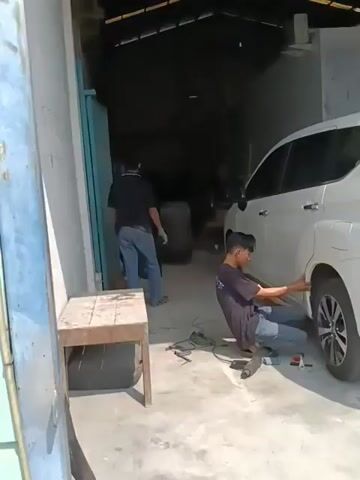 A man is working on the rear wheel of a white SUV, which is parked inside a garage. He is kneeling and appears to be using a tool to adjust something on the car's fender. Another man is standing in the background, seemingly working on a large tire. The garage has a concrete floor and an open entrance. Tools and car parts are scattered around the work area.