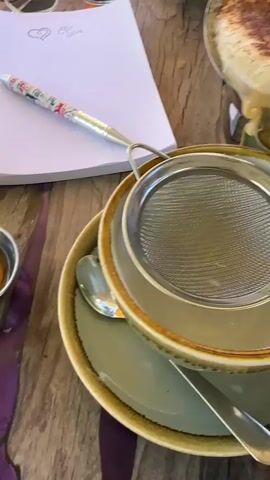 The video shows a close-up overhead view of a table setting. A white notebook with a pen and sunglasses is visible in the upper left quadrant. A small metal dish containing a golden liquid, possibly honey, is to the left of the notebook. In the center and right of the frame, a teacup sits on a saucer. A metal strainer is placed over the teacup, and a pot is being tilted to pour a frothy, light brown liquid through the strainer into the cup. The liquid contains visible solid particles, likely tea leaves or spices, which are caught by the strainer. The table surface appears to be made of wood with a textured, possibly painted or stained, finish.