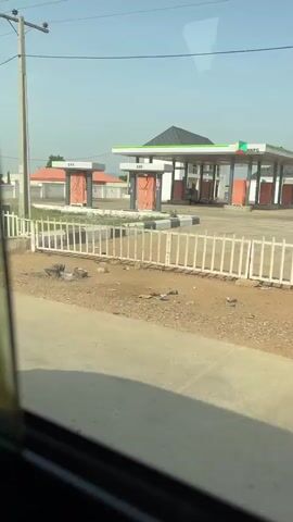 The video captures a continuous panning view from a moving vehicle, showcasing various roadside scenes under a clear, sunny sky.

The initial shot reveals a gas station with a green and white sign displaying "NIPCO," a Nigerian fuel retailer. Fuel pumps are visible with labels "DPK" and "AGO." A white picket fence separates the road from the station, which features several buildings, some with red roofs, and power lines overhead.

As the vehicle continues, the view transitions past a white building with a dark-tiled roof and a water storage tank on a stand. The scene then opens up to a wide, grassy area with scattered patches of dirt. In the background, there are several single-story buildings, including one with a red roof and another longer, yellow-hued structure, suggesting a school or institutional building.

The final segment of the video shows a commercial area along the road. Three young men are seen walking casually alongside a metal fence. Behind the fence, several compact pickup trucks, both white and red, are parked, possibly indicating a vehicle dealership or repair shop. Further in the background, more buildings, some appearing older or partially constructed, are visible. The overall setting suggests a semi-urban or peri-urban environment in Masaka, Nigeria.