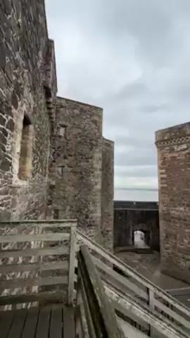 The video shows a stone castle from a high vantage point on a cloudy day. The camera pans from a wooden staircase between two castle towers to a stone walkway that leads to the edge of the castle overlooking the sea. The sea is visible in the distance, and there is a grassy area below the castle walls.