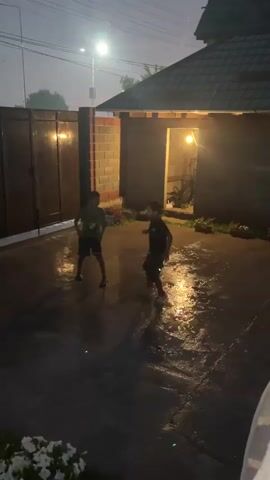 This video clip captures a group of four children playing outside on a wet, paved surface at dusk. The setting appears to be a residential backyard or courtyard, indicated by a dark metal gate on the left, a brick wall with an open doorway on the right, and a tiled roof structure. A streetlamp casts a bright glow from above, illuminating the scene and creating reflections on the wet ground. The sky is a dusky grey, suggesting either early morning or late evening. The ground is covered in puddles, and water appears to be splashing as the children move, implying it might be raining or they are actively playing with water.

The children, appearing to be between the ages of 6 and 10, are engaged in energetic movement. They are dressed in casual clothing, including t-shirts and shorts. Two children are in the foreground, and two are further back, with one child on the far right largely out of frame. Their body language suggests playfulness and excitement, with some jumping and others raising their arms. The overall atmosphere is one of carefree fun despite the wet conditions. There are no visible text elements in the clip.