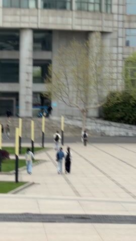 The video shows an exterior scene on what appears to be a university campus. Several individuals walk across a paved plaza in front of a large, multi-story building with a stone or concrete facade. The people are dressed in casual clothing, some carrying bags or musical instruments. There are also some yellow lampposts and patches of green grass in the foreground. A tree with bare branches stands near the building entrance. A few scooters are parked in the background. The camera appears to be positioned slightly elevated.