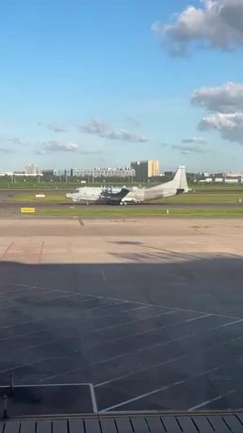 A large, white airplane with four propellers is on a runway at an airport. The plane is facing left and appears to be taxiing or preparing for takeoff. In the background, there are buildings and a blue sky with scattered clouds. The foreground shows a portion of an airport tarmac with various markings.