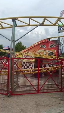 The video shows a roller coaster ride at an amusement park. The ride features a race car theme, with checkered flags and images of sports cars decorating the attraction. The roller coaster cars are red and have racing lights. The track is yellow and yellow support beams can be seen. In the background, trees and a cloudy sky are visible. The video is shot from a ground-level perspective, looking up at the roller coaster.