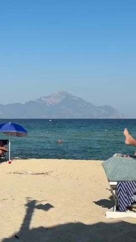 The video opens with a view of a sandy beach with turquoise water and a large mountain in the background. There are a few beach umbrellas, lounge chairs, and people relaxing on the beach. Some people are in the water swimming. The scene is bright and sunny, suggesting a warm day at the beach. The camera pans across the beach, showing more people enjoying the scenery and activities.