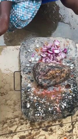 A pair of hands is shown mashing onions with a stone on a rough, rectangular stone slab. The onions are sliced and piled in the center of the slab, with some red paste and debris around them. The stone slab has a black metal handle on the left side and appears to be placed on a tiled floor. The hands are actively crushing the onions with the stone.