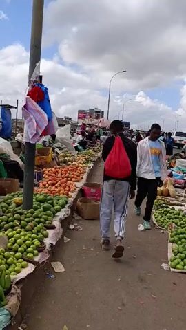 The video shows a busy open-air market under a partly cloudy sky. Rows of vendors sit behind displays of produce, including avocados, tomatoes, and other fruits and vegetables. People walk through the market, some carrying bags, browsing the goods for sale. The market appears to be on or adjacent to a bridge or overpass, with vehicles visible in the background.