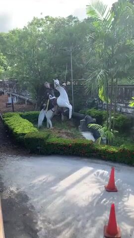 A statue of a person on a horse is shown. The statue is white and appears to be made of plaster or similar material. The person is wearing a helmet and dark clothing. The horse is also white. The statue is positioned on top of a large black pipe that is supported by concrete blocks. There are green bushes and trees in the background, as well as a concrete wall. A speed limit sign that reads "SPEED 15" is visible in the foreground. The video pans and zooms in on the statue, providing different angles.