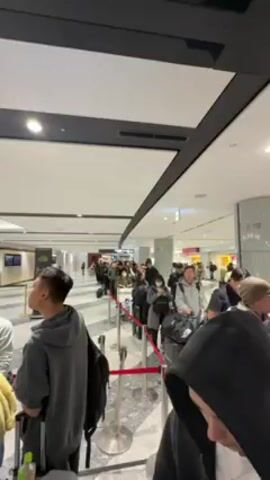 The video shows a queue of people in what appears to be an airport terminal. The people are diverse in age and attire, many with luggage, including backpacks and rolling suitcases. They are standing in a line marked by red velvet ropes and metal stanchions. The terminal is well-lit with a modern design, featuring a high white ceiling, recessed lighting, and gray marble-like flooring. In the background, there are shops and signage in what looks like a duty-free area, including a Coca-Cola display. The camera moves slightly as it films the scene, giving the impression of someone standing in the queue.