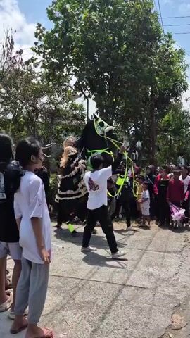 A group of people are gathered on a street, watching a performer dressed as a horse. The performer is black and green and adorned with colorful tassels. Another person, wearing a white t-shirt and black pants, controls the performer's movement with reins. The crowd consists of adults and children, some of whom are holding flowers. The background shows trees and houses under a clear sky.