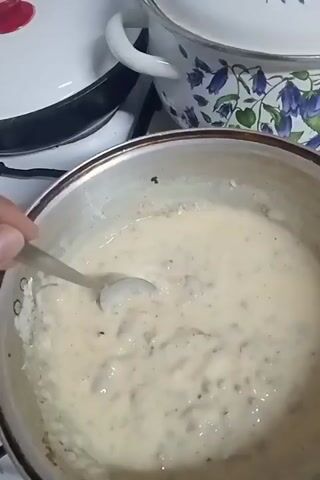 A close-up, overhead shot shows a metal pot on a stovetop, containing a creamy, light-colored substance that appears to be simmering. A hand is holding a spoon and stirring the contents of the pot. In the background, there are other pots and kitchen appliances visible. The lighting is dim, suggesting an indoor setting.