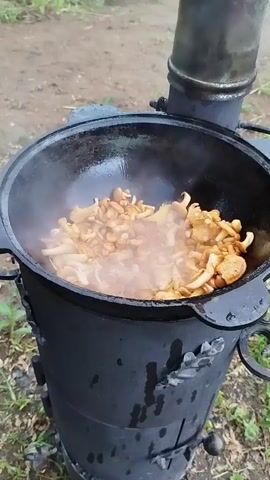 The video shows a close-up view of a black cast-iron pot filled with chanterelle mushrooms being cooked outdoors. The pot is heated by a black metal stove, and steam is rising from the mushrooms, indicating they are actively cooking. The mushrooms appear to be golden-yellow and are mixed with some spices or seasoning. The background is blurred but suggests an outdoor setting with dirt and some vegetation. The camera angle is slightly above the pot, looking down into it.