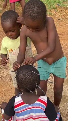 This media file features three young Black children in an outdoor setting with dirt ground and some greenery in the background, suggesting a rural or less developed area, possibly Achara, Nigeria. The central activity involves two children styling the hair of a third child.

The child in the foreground, facing away from the camera, has their hair being styled with what appear to be thin, dark strands, possibly wire or thick thread, which are being inserted into their hair, giving it a spiky appearance. The child in the middle ground, a shirtless boy wearing teal shorts, is actively engaged in this process, using his hands to weave or insert the material into the other child's hair. Another child, wearing a yellow shirt, is partially visible on the left, also seemingly involved or observing the activity.

The lighting suggests it is daytime, with no strong indicators of specific time of day or weather conditions beyond it being outdoors. The children appear focused on their activity, though their emotions are not clearly discernible from the image. There is no visible text within the frame of this media file.