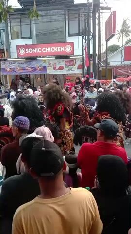 The video captures a street scene with a crowd of people gathered to watch a performance. In the foreground, several individuals are seen from behind, some wearing hats and casual clothing. The performers, dressed in elaborate costumes with large, dark, and voluminous hairpieces, are the focus of the event. They appear to be dancing or moving in a procession. Behind the performers, a street is lined with various shops, including "SONIC CHICKEN" and "MOMOYO ICE CREAM," indicating an urban or semi-urban setting. Banners and signs are visible, suggesting a festive or promotional event. The atmosphere seems lively and engaging, with many people holding up phones to record the spectacle.