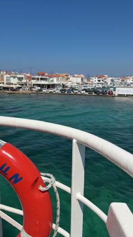 A first-person perspective shot from a boat reveals a bright blue sky and a calm, turquoise ocean. The foreground features the white railing of the boat and a red life preserver. In the middle ground, a coastal town with white buildings and parked cars is visible. The background shows distant hills under the clear sky. The boat appears to be moving slowly through the water.