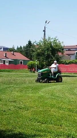 A person wearing a white hat and a white shirt is riding a green John Deere lawn tractor. The tractor is moving across a grassy field. In the background, there are houses with red roofs and green fences, as well as trees. The sky is clear and blue. The sun is shining, casting shadows on the grass. The person appears to be mowing the lawn.