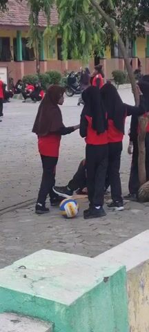 A group of children in red shirts and black pants are playing volleyball on a paved schoolyard. One boy in the foreground is about to serve the ball, while others are positioned to receive it. In the background, more children are visible, some observing the game and others walking around. The school building is visible behind the children, with windows and colorful decorations.