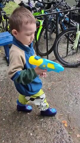 A young boy, likely under the age of four, stands on a wet asphalt surface holding a blue and yellow water gun. He is wearing a blue and tan fleece jacket over a dark green long-sleeved shirt, green pants with a white character print, and dark blue rain boots. The boy is looking directly at the camera with a neutral expression.

Behind him, several bicycles are parked. A white car with a license plate reading "C933CE 72" is visible in the background. The ground is wet, suggesting it has recently rained or is raining. The setting appears to be an outdoor area, possibly a street or park, with bicycles parked nearby. The lighting suggests it is daytime. The overall mood is casual and depicts a child ready to play.