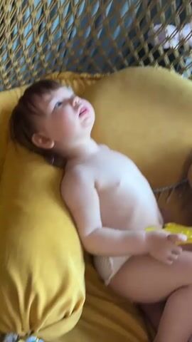 A young child lies in a hammock. The child is fair-skinned with brown hair, wearing only a diaper. The child lies on a yellow cushion, looking upward with an open mouth. The hammock is made of woven rope with blue backing. The child holds a yellow object in their right hand. The room is bathed in soft, indirect sunlight.