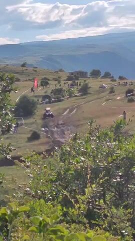 This media file captures an off-road buggy race event taking place in Antarut, Armenia. The scene is set on a hilly, mountainous terrain with a clear blue sky and scattered clouds, suggesting it's daytime. The event appears to be well-attended, with spectators lining parts of the racecourse.

The primary subject is a dark-colored off-road buggy, kicking up dust and mud as it navigates a dirt track. The buggy is positioned in the middle of the frame, moving from left to right and slightly away from the viewer. Around the track, several people are visible, some appearing to be spectators, while others might be event staff. There are flags and banners, including a prominent red flag with white lettering, marking the course.

The landscape features rolling hills covered in green grass and some scattered trees and bushes, particularly in the foreground, which are lush and green. In the background, more distant hills and valleys are visible, suggesting a vast, open natural setting. A few buildings are sparsely located on the hills in the distance, hinting at a rural or remote location. The overall atmosphere seems energetic, typical of a sporting event.

There is a visible white banner with red lettering, and a red flag with white graphics. The activity is clearly a race, with the buggy in motion and people gathered to watch. The weather appears to be fair, suitable for an outdoor event. The combination of the rugged terrain and the racing buggy creates a dynamic and adventurous scene.