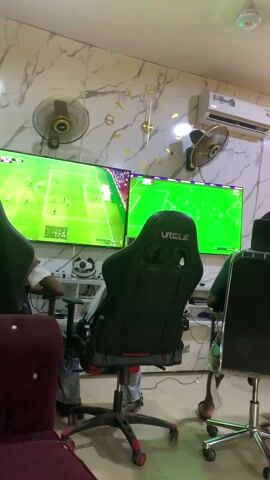 In a small room with marble-patterned walls, two people are playing a video game on two large televisions. Each player sits in a gaming chair, one labeled “PURPLE”. The game appears to be a soccer simulation, with green fields displayed on the screens. There are electric fans mounted on the wall above each television. A decorative gold clock with Roman numerals is visible on the wall between the screens. An air conditioner is installed high on the wall. One of the players is wearing a white shirt. A decorative soccer ball helmet sits on a shelf below one of the screens.