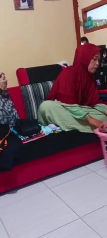 A woman wearing a maroon hijab and a light green shirt is sitting on a red sofa. She is folding clothes, holding a dark blue shirt with red accents in her hands. In front of her, on the sofa, is a pile of folded clothes. To her left, there is a black bag and more folded clothes. To her right, a pink laundry basket filled with clothes is visible. In the background, a man is standing near a shelf with electronics and jars. The room has yellow walls and a tiled floor. The woman is actively engaged in folding clothes.