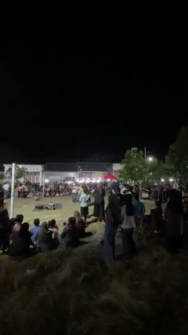 The video shows a crowd of people gathered at night, likely for an outdoor event. In the foreground, a group of people are seated on the ground, facing away from the camera. In the middle ground, more people stand, some observing a performance or activity taking place on a stage in the background. The stage is illuminated with red lights and features a large screen. There are buildings and trees visible in the background, with lights suggesting a populated area. The overall atmosphere appears to be festive and communal.