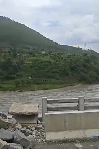 The video shows a wide, muddy river flowing through a valley with steep, green, forested hillsides. A modern concrete bridge with grey railings is in the foreground, obscuring part of the view. The sky is overcast with dramatic clouds. In the distance, a thin suspension bridge spans the river. The overall impression is one of a remote, natural landscape with man-made infrastructure.