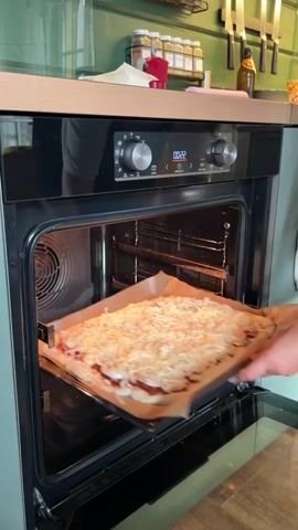 A person's hand is shown placing a baking tray with a rectangular pizza topped with cheese into an open oven. The oven has a digital display showing the time as 00:22 and control knobs. The pizza is on parchment paper and appears to have a red sauce base. The oven interior is lit, and rack guides are visible. The kitchen environment is suggested by a countertop and cabinetry in the background. The camera angle is focused on the oven opening and the pizza being inserted.