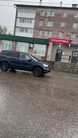 The video depicts a dark blue SUV, likely a Honda CR-V (3rd generation), on a wet asphalt surface in what appears to be an urban or semi-urban setting in Mozhga, Russia, on an overcast, rainy day. At the beginning of the video, the front right wheel of the SUV is significantly elevated, suggesting it is stuck or balancing precariously on uneven terrain or in a pothole filled with water. The vehicle then slowly moves forward and drives away.

In the background, there are brick buildings with multiple windows, some of which have air conditioning units installed. One building features a green roof. On the ground floor of one building, a red sign with white text is visible, reading "ГОСАПТЕКА" (GOSAPTEKA), which translates to "State Pharmacy." Power lines stretch across the frame above the buildings. The ground is visibly wet with puddles, indicating ongoing or recent rainfall. Trees are visible in the distance between buildings.