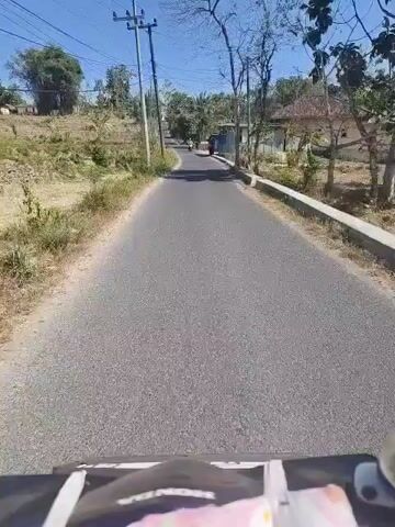 The video shows a point-of-view shot from a vehicle moving along a paved road. The road is flanked by dry grass and sparse vegetation on the left, and houses and trees on the right. In the distance, the road curves slightly uphill. A motorcycle with a rider is seen approaching from the opposite direction. The sky is clear and blue, indicating a sunny day.