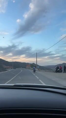 The video is filmed from inside a car, capturing a road trip.  A motorcyclist rides ahead on the road, moving from left to right, passing a turnoff and road signs.  The background features mountains beneath a sky with colorful sunset tones. Power lines are visible alongside the road. The car is moving along the road.