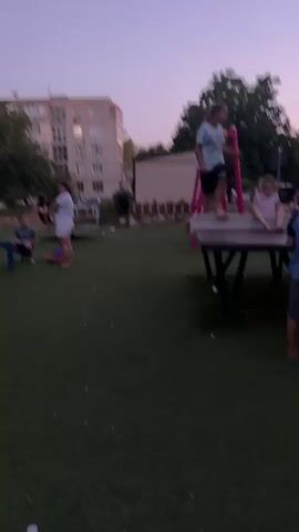 The video shows a playground at dusk. There are several children and at least one adult present. In the foreground, a young girl in a blue and white striped shirt is standing on a piece of playground equipment, possibly a balance beam or a raised platform. She appears to be in motion, possibly jumping or balancing. To her left, another young girl in a pink top is sitting on a similar piece of equipment. In the middle ground, a woman in a white t-shirt and shorts stands looking at her phone, which she holds up to take a picture or video. A boy in a blue shirt is sitting on the ground near her. Further in the background, there is a larger playground structure with slides and other colorful equipment, and a few more children can be seen playing. An apartment building and trees are visible in the background. The ground is covered with green artificial turf. The lighting suggests it is early evening.