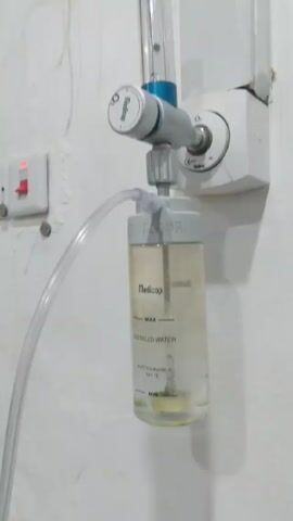 The video shows a medical oxygen flow meter and humidifier attached to a wall. The flow meter is mounted on a white wall outlet. Connected to the flow meter is a clear plastic humidifier bottle labeled "Medicop". The bottle has markings for "MAX," "DISTILLED WATER," and "MIN." The water level in the bottle is between the "MIN" and "MAX" markings. A clear plastic tube extends from the humidifier bottle. Above the flow meter, a vertical tube with a blue top appears to indicate the oxygen flow rate. To the left of the flow meter and humidifier, there is a light switch on the wall. The wall is painted white and has a slightly textured surface. The lighting is relatively dim.