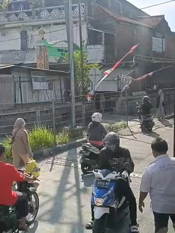 The video shows a street scene with several motorcycles and people. In the foreground, a person on a blue and white motorcycle with the license plate 2252 RN is riding. To the left, a motorcycle with a yellow basket and what appears to be a metal box is also present. Behind these, other motorcycles and individuals are visible, some of whom are wearing headscarves. The background features buildings, including one with what looks like a mosque or religious structure, and some fences. The overall environment suggests a bustling street in what could be an urban or semi-urban area. The lighting indicates it is daytime, likely sunny.