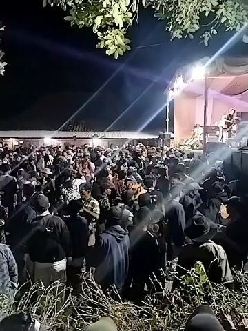 The video shows an outdoor nighttime event with a live band performing on a stage. A large crowd of people is gathered in front of the stage, watching the performance. The area is illuminated by bright stage lights and spotlights, creating dramatic lighting effects. The audience appears to be actively engaged, with many people standing and some possibly dancing. The environment suggests a casual gathering, perhaps a concert or festival, with trees and foliage visible in the foreground and background, indicating an outdoor setting. The crowd is diverse in terms of clothing and apparent age. The overall atmosphere is energetic and lively.