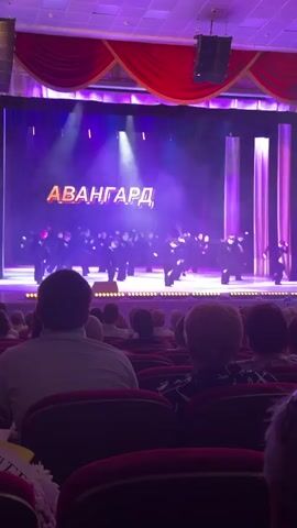 A mid-angle, medium shot shows a stage performance from the perspective of an audience member. On the stage, a group of dancers in dark costumes performs in front of a large screen displaying the word "АВАНГАРД" (AVANGARD) in orange. The stage is brightly lit with purple lights and has red curtains on the sides. In the foreground, the backs of several audience members' heads are visible as they watch the performance. The audience members are seated in red theater seats. The shot is stable, and the lighting is consistent.