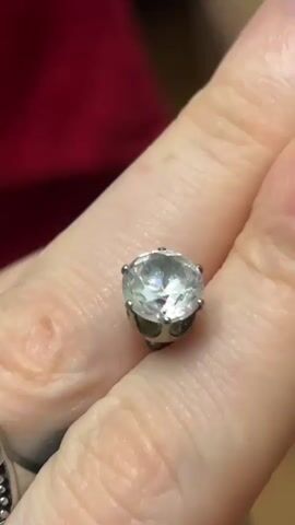 A close-up shot shows a person's hand holding a ring with a large, clear, faceted gemstone set in a silver-toned metal band. The gemstone appears to be a cubic zirconia or a diamond. The ring is held against a blurred background of a red garment. The person's fingers are visible, with some visible fingernails and skin texture. Another ring with a patterned band is partially visible on a lower finger. The lighting is bright, highlighting the sparkle of the gemstone.