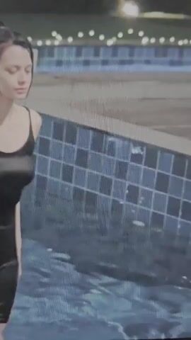 The video begins with a close-up of a young woman in a black swimsuit, kneeling in a pool. Her hair is wet and slicked back. The pool has blue and black tiled edges. In the background, string lights are visible. The camera then transitions to a closer shot of the woman's face and upper body. She is wearing a black, sheer robe over a black lace slip. Her hair is still wet and hangs loosely around her shoulders. She looks down with a somber expression. The scene appears to be outdoors at dusk or night.