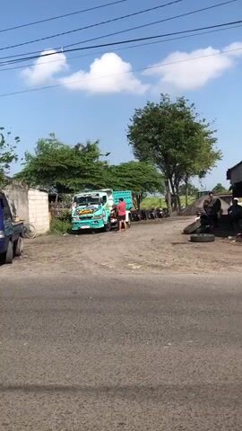 The video shows a blue pickup truck driving past a road with a truck and some people standing around. The truck is parked in front of a building and there are trees in the background. The sky is blue with some clouds. The video is shot from the perspective of someone standing on the side of the road. The blue pickup truck appears to be a Suzuki Carry pickup.