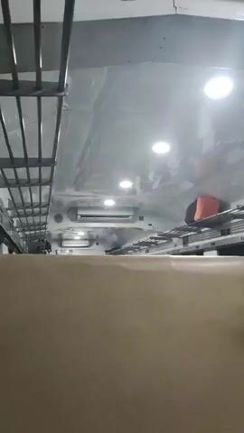 The video shows the interior of a train carriage. The camera pans from right to left, capturing the ceiling with multiple lights, an air conditioning unit, and luggage racks above seats. A few pieces of luggage, including an orange bag, are visible on the racks. The seats themselves are covered in a light brown fabric. The overall environment appears clean and functional, consistent with typical train travel. There are no people clearly visible in the frame, and no specific actions or interactions are depicted. The lighting suggests it may be daytime or the interior lights are on.