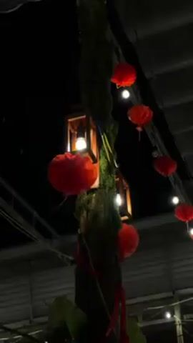 A video captures a decorative scene at night. A tall, moss-covered post stands prominently, adorned with wooden lanterns housing glowing light bulbs and red, paper-like lanterns scattered around it. Ribbons in red, blue, and yellow are also attached to the post. The background is a dark, open space with a metal framework visible above, punctuated by string lights. Sounds of chatter and children's voices can be heard in the background.