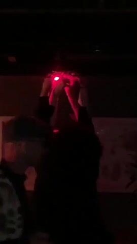 The video shows a person in a dark environment, possibly a bar, holding a small red light source above their head. The red light illuminates their face and hands. The person is wearing a dark shirt. There is a framed picture on the wall behind them, but the details are indistinct. Another person, partially visible in the foreground, has a pattern on their jacket or shirt.