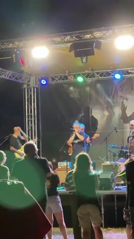 A live band performs on an outdoor stage at night. The stage is lit with bright overhead lights and some colored lights. A lead singer in the center of the stage is playing a harmonica and singing into a microphone. To his left, a guitarist is playing a guitar. To his right, a bassist is playing a bass guitar. A drummer is partially visible behind the bassist. A crowd of people are standing in front of the stage, watching the performance. Some of the audience members are silhouetted against the stage lights. The background behind the band is a dark curtain.